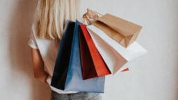 A person with long blonde hair holds several colorful paper shopping bags over their shoulder against a plain background.