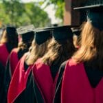 A group of graduates in caps and gowns with red stoles walk outdoors in a line on a sunny day.