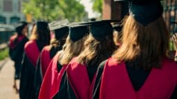 A group of graduates in caps and gowns with red stoles walk outdoors in a line on a sunny day.