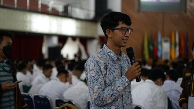 A man in a patterned shirt speaks into a microphone at an indoor event with a seated audience in the background.