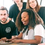 Five people looking at a laptop screen together, appearing engaged and focused in a bright office setting.