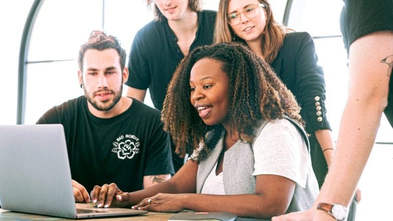 Five people looking at a laptop screen together, appearing engaged and focused in a bright office setting.