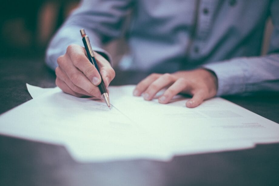 A person wearing a blue shirt writes on papers at a desk with a pen.