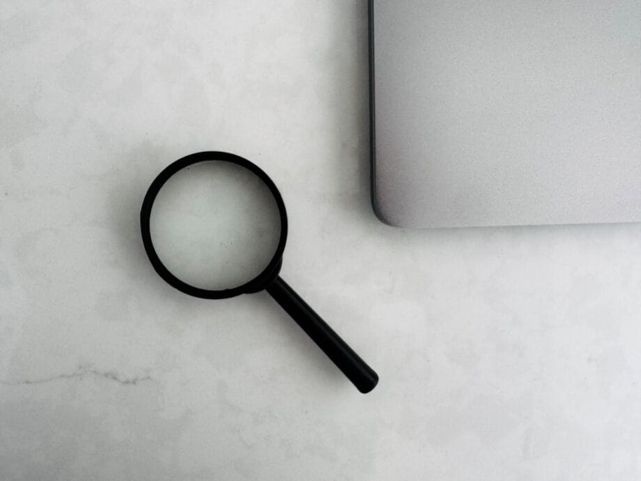 A magnifying glass lies next to the corner of a closed silver laptop on a white surface.