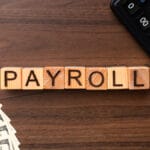Wooden blocks spelling PAYROLL on a desk with a fan of dollar bills and part of a calculator visible.