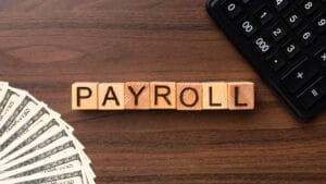 Wooden blocks spelling PAYROLL on a desk with a fan of dollar bills and part of a calculator visible.