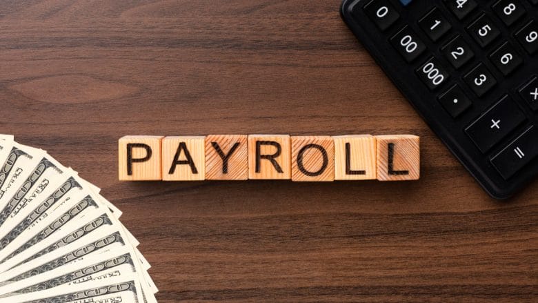 Wooden blocks spelling PAYROLL on a desk with a fan of dollar bills and part of a calculator visible.