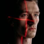 A man’s face is illuminated by intersecting red laser lines in a dark setting.