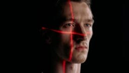 A man’s face is illuminated by intersecting red laser lines in a dark setting.