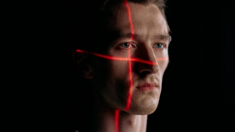 A man’s face is illuminated by intersecting red laser lines in a dark setting.