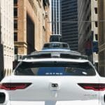 A white Waymo self-driving car with sensors on the roof drives through a city street surrounded by tall buildings.