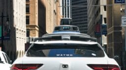 A white Waymo self-driving car with sensors on the roof drives through a city street surrounded by tall buildings.
