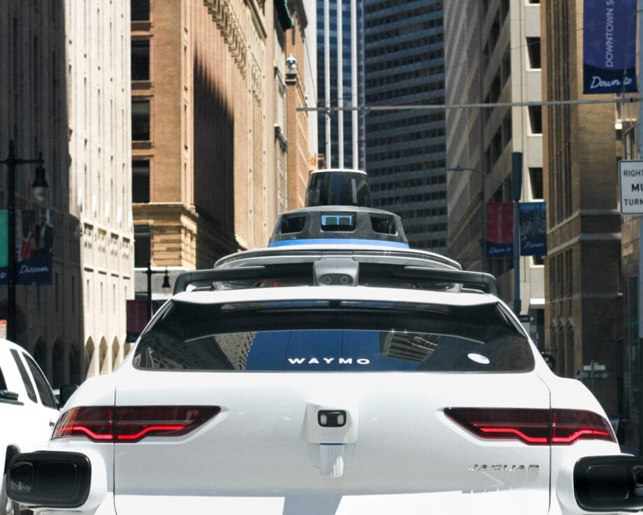 A white Waymo self-driving car with sensors on the roof drives through a city street surrounded by tall buildings.