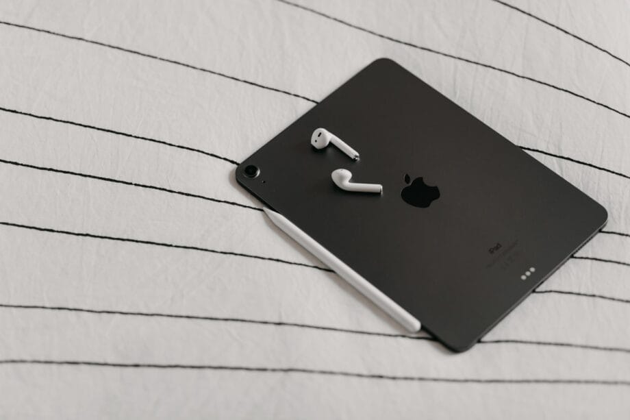 A black iPad and white stylus with wireless earbuds on top, resting on a white bed with black stripes.