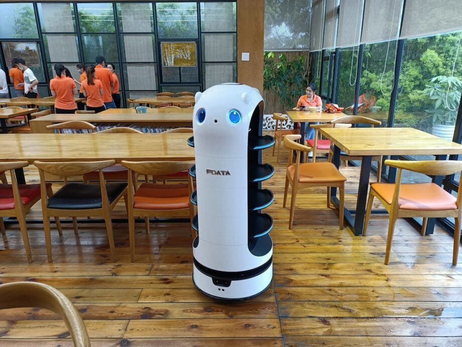 A white service robot with shelves stands in the middle of a restaurant with wooden tables and chairs.