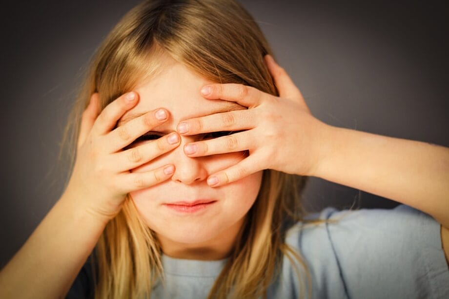 A young child covers their eyes with both hands while slightly smiling, standing against a plain background.