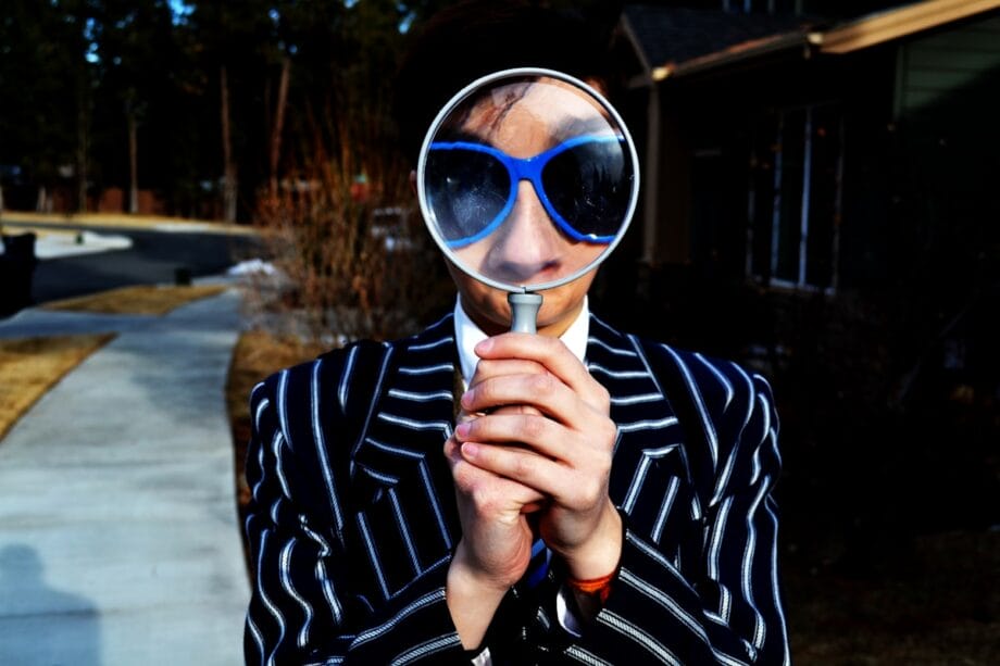 Person in a striped suit holding a magnifying glass up to their face, magnifying their sunglasses, standing outdoors on a sidewalk.