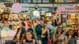 Crowd of people walking through an indoor market with various food stalls and signs visible in the background.