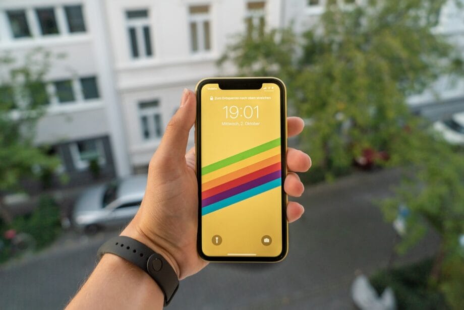 A hand holds a smartphone with a rainbow-striped wallpaper, displaying the time 19:01 and the date Wednesday, October 2.
