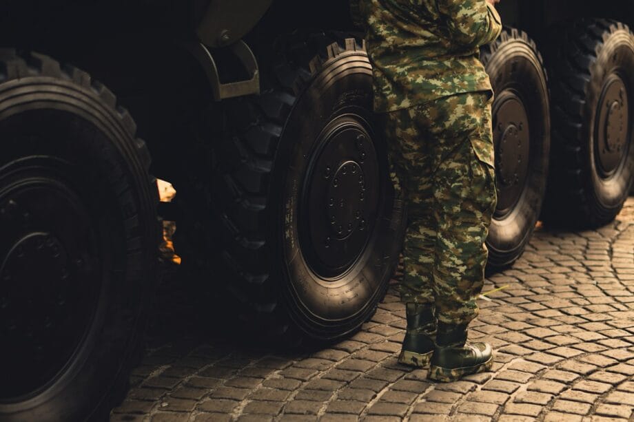 A person in camouflage military uniform stands next to large vehicle wheels on a cobblestone street.