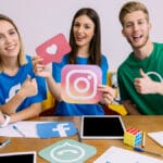 Three people at a table hold social media icons and give thumbs up, with laptops, tablets, and paperwork in front of them.