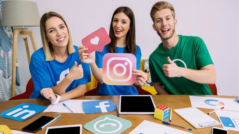Three people at a table hold social media icons and give thumbs up, with laptops, tablets, and paperwork in front of them.