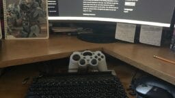 A desk with a retro-style keyboard, games controller, mouse, notebook with Halo cover, and a monitor displaying a website.