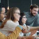 Four people sitting at a desk in an office, collaborating and looking at a computer screen together.