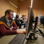 Several people wearing headphones work at desktop computers in a row inside a wood-paneled office or computer lab.