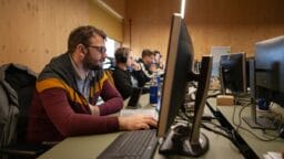 Several people wearing headphones work at desktop computers in a row inside a wood-paneled office or computer lab.