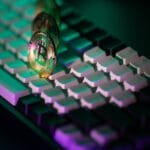 A Bitcoin coin stands upright on a black and white computer keyboard, illuminated by green and purple light.