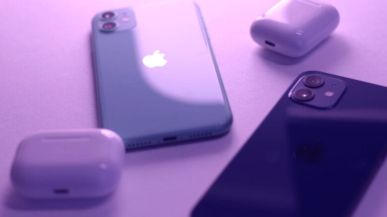 Two iPhones and two AirPods cases are placed on a flat surface under purple lighting.