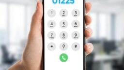A hand holding a smartphone displaying a dial pad with the number 01225 entered, in an office setting.