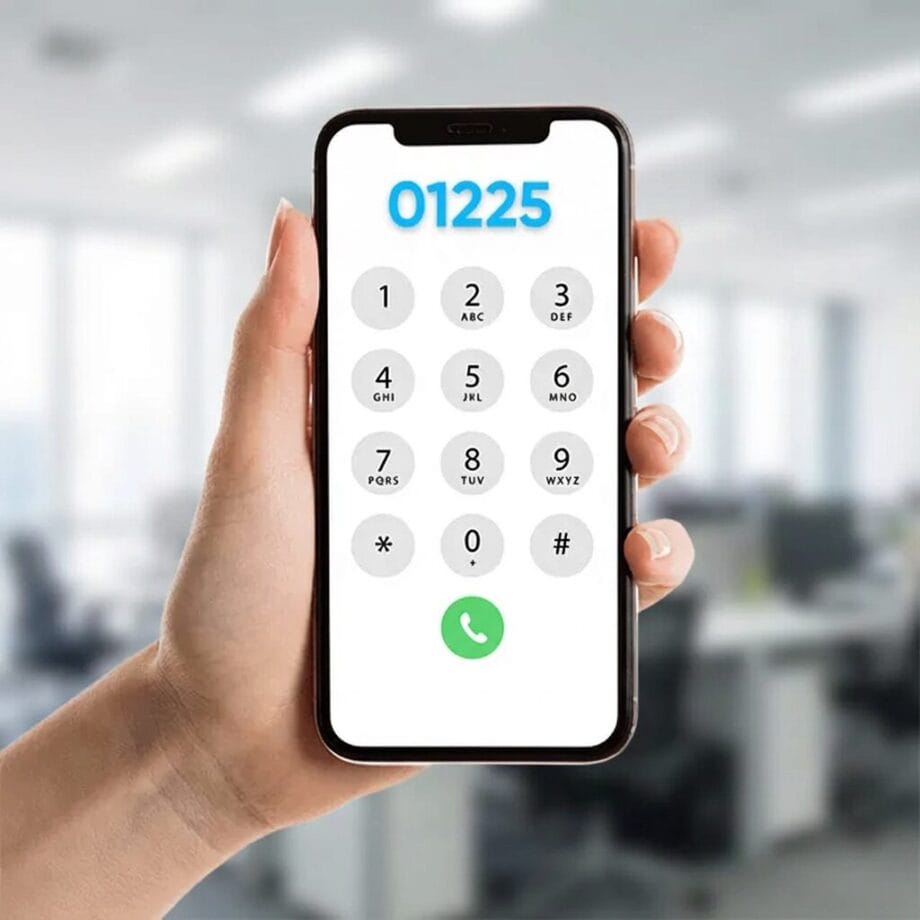 A hand holding a smartphone displaying a dial pad with the number 01225 entered, in an office setting.