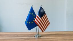 Two small flags, one of the European Union and one of the United States, stand side by side on a metal holder on a wooden table.