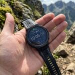 A hand holds a digital sports watch displaying the time and stats, against a rocky mountain landscape background.