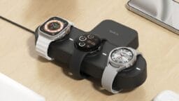 A charging station with two smartwatches and a smartphone on a wooden desk, alongside a cup and other small items.