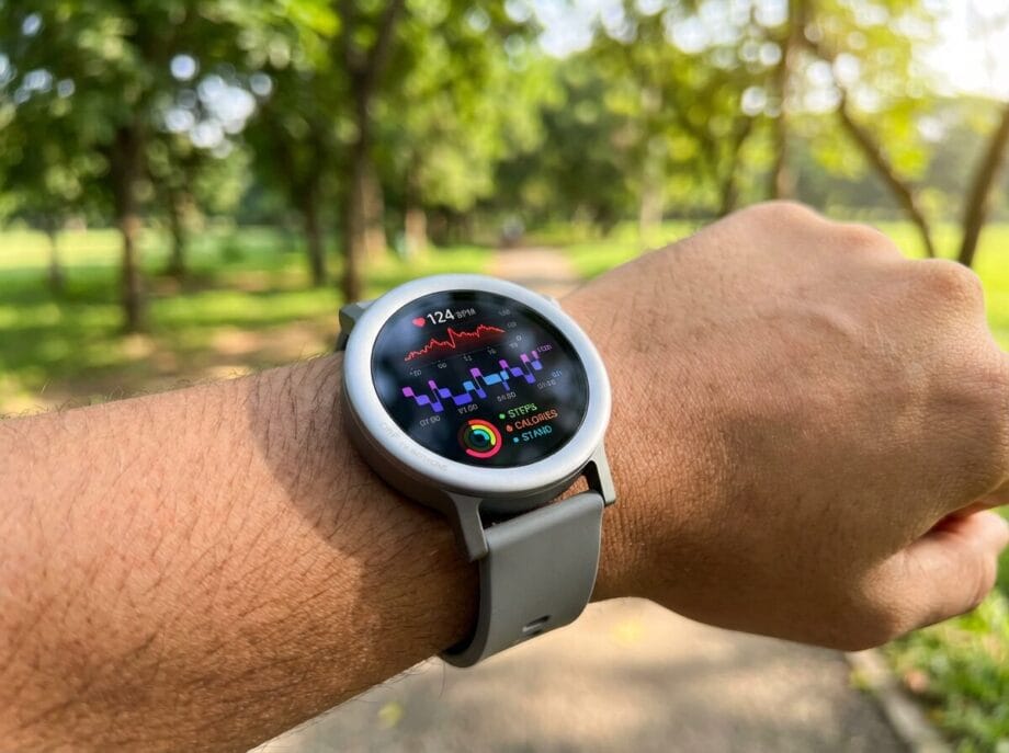 A person wearing a smartwatch displaying heart rate, activity rings, and step count while standing on a tree-lined path.