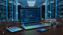 A laptop with code on the screen, tablet, smartphone, headphones, and a SOFTWARE sign on a desk in a server room.