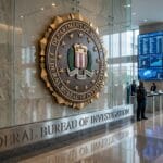 FBI headquarters lobby with the agency seal on the wall, people walking, and monitors displaying information; U.S. Capitol visible outside.