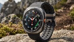 A rugged smartwatch with a black band and colorful dial is displayed outdoors on a rocky surface with mountains in the background.