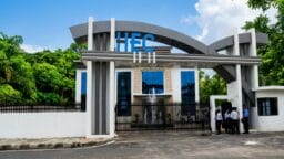 Modern building entrance with HEC sign, glass facade, and several people standing by the gate on a sunny day.