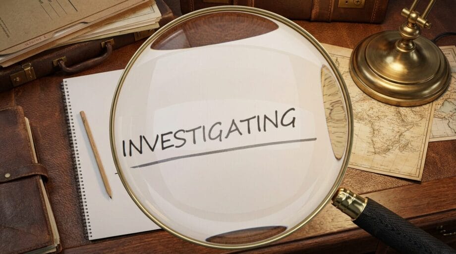 A magnifying glass enlarges the word INVESTIGATING written in a notebook on a desk with books, papers, and a map.