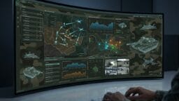 A person operates a computer displaying military maps, charts, and surveillance data on a large curved screen with camouflage patterns.