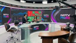 Two news anchors sit at a glass desk in a modern TV studio with NDTV Profit branding and stock market screens in the background.