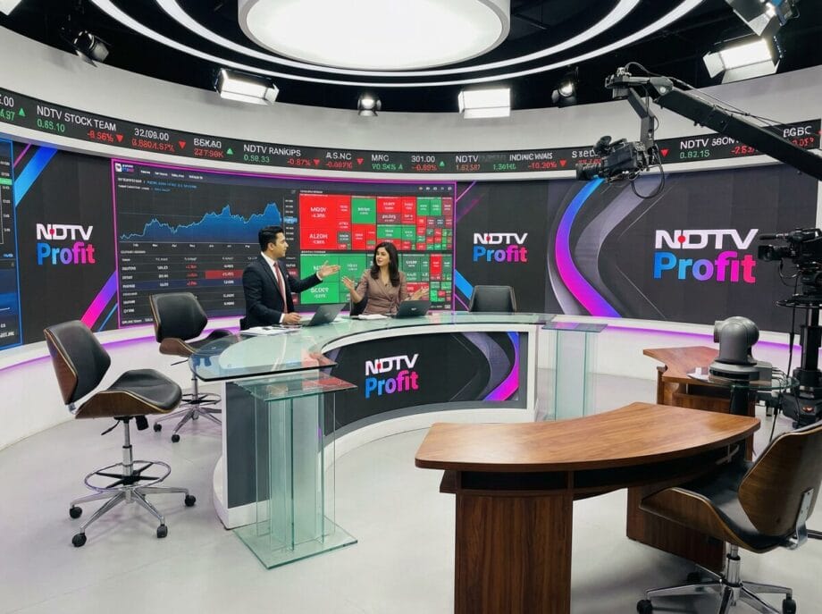 Two news anchors sit at a glass desk in a modern TV studio with NDTV Profit branding and stock market screens in the background.
