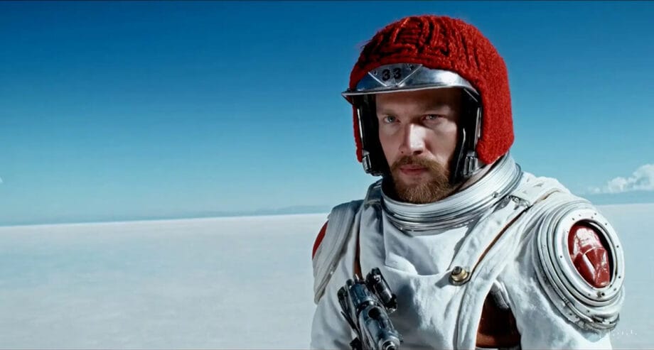 A person in a white spacesuit and red helmet stands on a flat, white landscape under a clear blue sky.