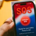 A hand holds a smartphone displaying an Emergency SOS screen with instructions to press to call for safety assistance.