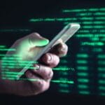 A hand holds a smartphone with green lines of code projected in the background, suggesting hacking or cybersecurity.