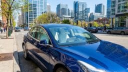 A blue electric car is parked on a city street with tall glass buildings and trees in the background.